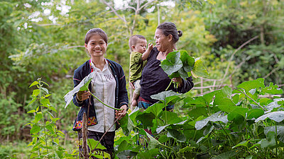 Zwei Frauen und ein Kleinkind stehen inmitten grüner Pflanzen. Die Frauen lächeln und halten große Blätter in der Hand. Im Hintergrund ist dichte Vegetation und Wald zu sehen.