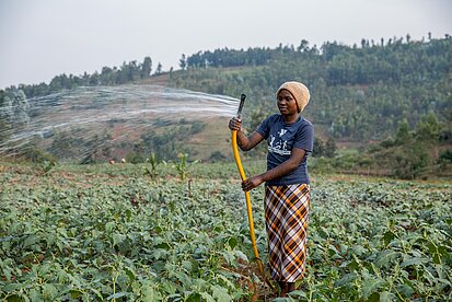 Eine junge Frau bewässert ihr Feld mit einem gelben Gartenschlauch. Sie trägt ein T-Shirt und einen karierten Rock. Im Hintergrund liegen grüne Hügel und Felder.