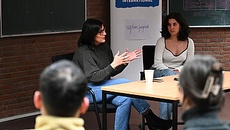 Maryam Blumenthal sitzt an einem Tisch in einem kleinen Hörsaal vor einem Plan-Aufsteller.