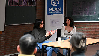 Maryam Blumenthal sitzt an einem Tisch in einem kleinen Hörsaal vor einem Plan-Aufsteller.