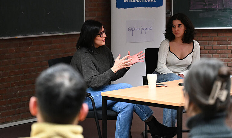 Maryam Blumenthal sitzt an einem Tisch in einem kleinen Hörsaal vor einem Plan-Aufsteller.