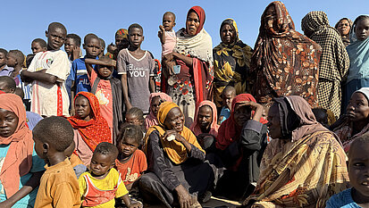 Eine Gruppe von Frauen und Kindern sitzt dicht beieinander im Freien auf sandigem Boden. Viele tragen farbenfrohe Kleidung und Kopftücher, einige blicken direkt in die Kamera, während andere miteinander sprechen.
