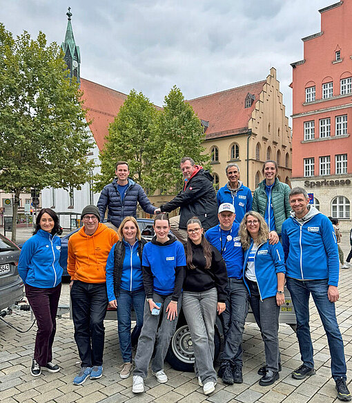 Eine Personengruppe in blauen Jacken mit dem Plan-Logo steht auf einem Platz zwischen historischen Häusern