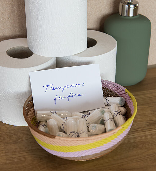 Korb mit Tampons Ein kleiner Korb mit Tampons, in dem ein Schild liegt, das auf Englisch erklärt, die Tampons wären kostenlos. Im Hintergrund sind Klorollen und ein grüner Seifenspender drapiert.