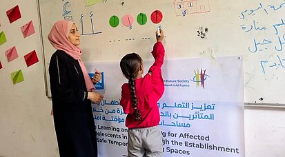 Amal steht mit einer Lehrerin vor einer Tafel und schreibt etwas auf.