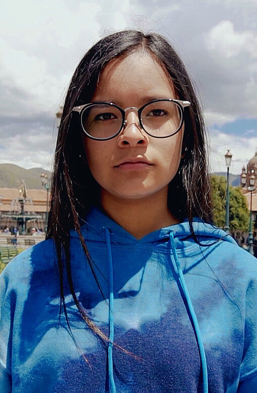 Astrid setzt sich in Cusco für sichere Räume für eingewanderte und geflüchtete Kinder ein. Astrid schaut ernst in die Kamera. Sie trägt eine Brille und einen blauen Pullover.