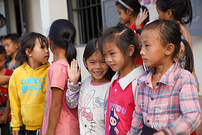 Eine Gruppe von Kindern steht nebeneinander vor einem Gebäude. Einige Mädchen lächeln und winken, andere schauen sich gegenseitig an. Die Kinder tragen farbenfrohe Kleidung und wirken fröhlich.