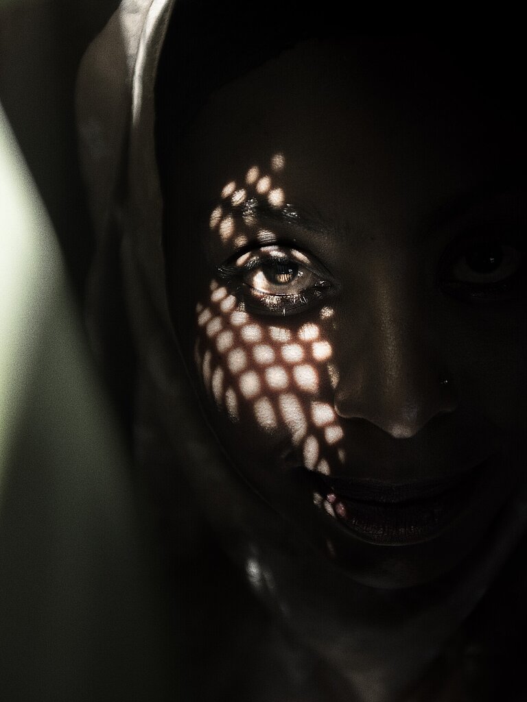 Ein Porträt einer Frau, auf deren Gesicht im Schatten ist. Ein wenig Licht scheint durch ein Gitter, das Muster des Schattens ist auf ihrem Gesicht zu sehen.