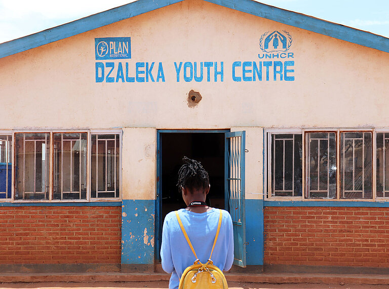 Clementine steht mit dem Rücken zur Kamera. Vor ihr ist ein Gebäude, auf dem Dzaleka Youth Centre steht. 