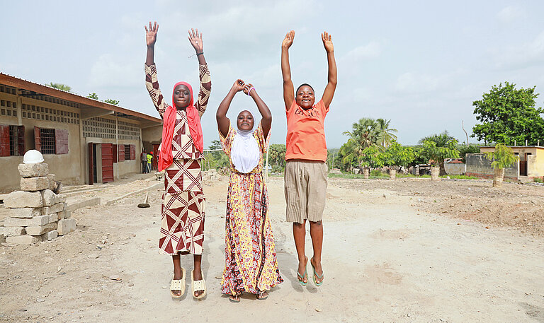 Drei Mädchen stehen auf einem sandigen Vorplatz einer Schule und springen in die Luft