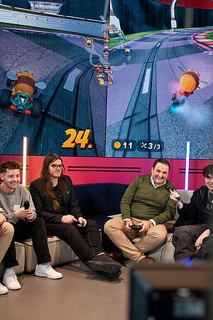 Vier Personen sitzen auf einem Sofa und spielen gemeinsam Videospiele, wahrscheinlich Mario Kart, auf einer großen Leinwand im Hintergrund. Eine Person hält ein Mikrofon, die anderen Controller. 