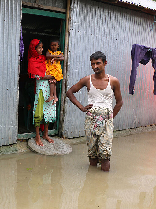 In Bangladesch wurden viele Regionen im Sommer 2022 überflutet. Eine Frau steht mit ihrem Kleinkind auf dem Arm in einem Hauseingang. Die Straße davor ist überflutet, ein Mann steht knietief im Wasser.