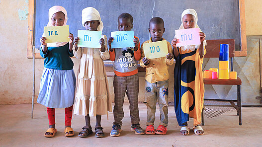 Fünf Grundschulkinder stehen nebeneinander vor einer Tafel und halten Schilder in der Hand.