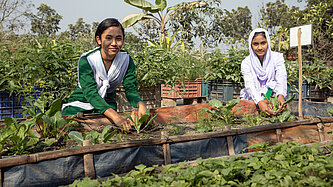 Zwei Mädchen in grüner Schuluniform hocken hinter einem Beet und lächeln in die Kamera