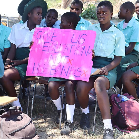 Eine Gruppe von Schüler:innen sitzt auf Stühlen und hört zu. Eine Schülerin hält ein leuchtend pinkes Plakat, auf dem steht: "GIVE US EDUCATION, NOT HUSBANDS". Das Plakat ist in großen, bunten Buchstaben geschrieben. Die Schüler:innen tragen Schuluniformen. 