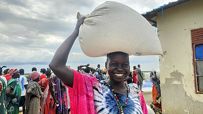 Eine junge Frau trägt lächelnd einen großen Sack mit Lebensmitteln auf dem Kopf. Im Hintergrund stehen weitere Menschen in einer Schlange, offenbar bei einer Verteilung von Hilfsgütern.