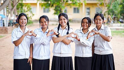 Fünf Mädchen aus Kambodscha im Teenager-Alter stehen in Schuluniformen vor ihrer Schule. Sie lächeln und formen mit ihren Händen Herzen.