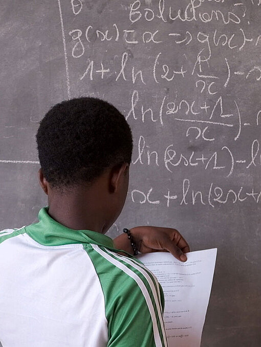 Absetou geht inzwischen wieder zur Schule. Absetou steht vor einer Tafel mit mathematischen Formeln und schaut auf ihr Arbeitsblatt.