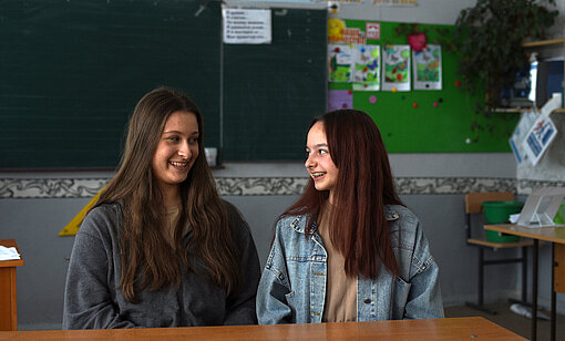 Zwei Mädchen sitzen in einem Klassenzimmer und schauen sich lächelnd an