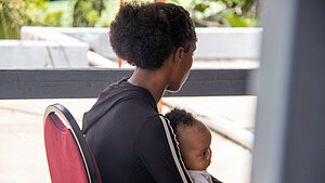 Eine Frau sitzt auf einem Stuhl und hat ein Baby auf dem Schoß. Sie sind von der Seite zu sehen. Die Frau schaut weg.