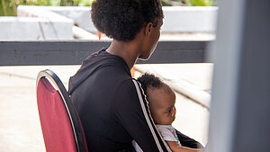 Eine Frau sitzt auf einem Stuhl und hat ein Baby auf dem Schoß. Sie sind von der Seite zu sehen. Die Frau schaut weg.