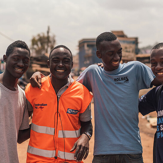 Vier junge Männer stehen Arm in Arm auf einer Straße und lachen in die Kamera.