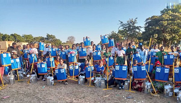 Eine große Gruppe von Menschen sitzt und steht im Freien und hält blaue Rückensprühgeräte sowie Metallkannen. Die Geräte sind vor den Personen aufgereiht, im Hintergrund sind Bäume und offene Fläche zu sehen.