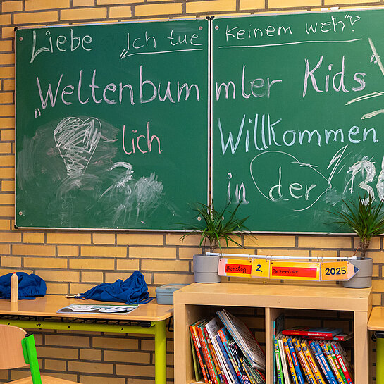 Klassenraum einer Grundschule mit Tafelaufschrift „Weltenbummler Kids – Willkommen in der 3c“