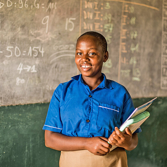 Ein Mädchen steht mit Schulmappe und Lerncomputer vor einer Tafel.