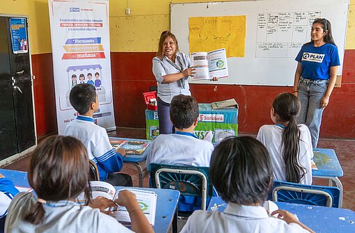 Mit Trainingsangeboten für wirtschaftliche und berufliche Eigeninitiativen engagiert sich Plan International an insgesamt 192 öffentlichen Schulen in Peru Eine Gruppe Schulkinder schaut zur Tafel, vor der eine Lehrerin mit einem aufgeklappten Buch steht