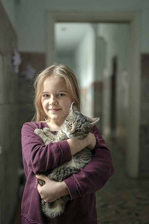 Ein Mädchen mit langen blonden Haaren hält eine grau-braun getigerte Katze auf dem Arm