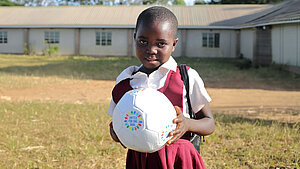 Ein Mädchen steht auf einem sonnigen Schulhof oder Sportplatz und trägt eine Schuluniform aus weißem Hemd und bordeauxfarbenem Kleid. Sie hält einen weißen Fußball in den Händen, auf dem das Logo „United for One Goal“ mit bunten Sternen zu sehen ist. Im Hintergrund steht ein Schulgebäude.