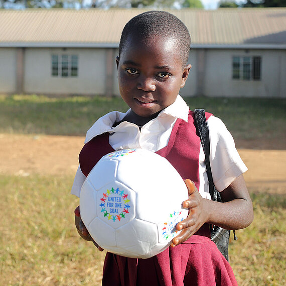Ein Mädchen steht auf einem sonnigen Schulhof oder Sportplatz und trägt eine Schuluniform aus weißem Hemd und bordeauxfarbenem Kleid. Sie hält einen weißen Fußball in den Händen, auf dem das Logo „United for One Goal“ mit bunten Sternen zu sehen ist. Im Hintergrund steht ein Schulgebäude. 