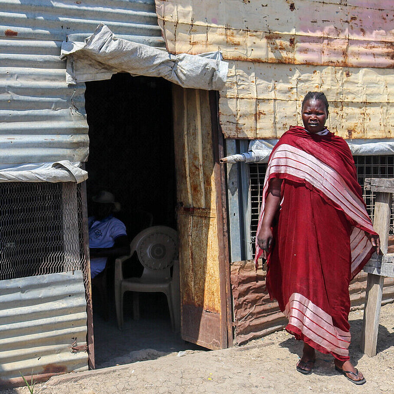 Vor einer Wellblechhütte steht eine Frau und lächelt