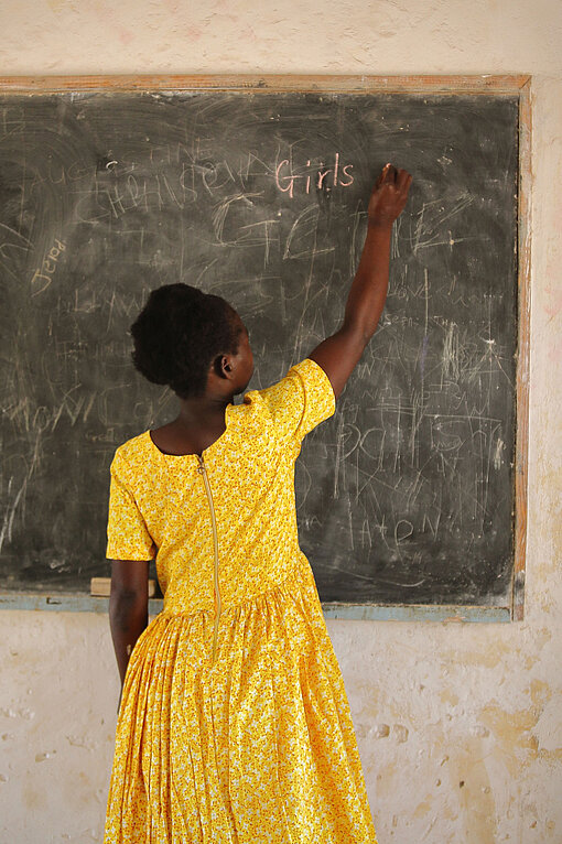 Ein Mädchen in einem strahlend gelben Kleid steht mit dem Rücken zur Kamera und schreibt das Wort "Girl" an eine Tafel.