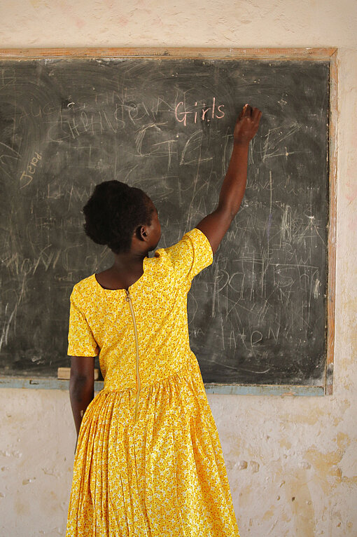Plan International verfolgt den Ansatz der kindorientierten Gemeindeentwicklung. Ein Mädchen in einem strahlend gelben Kleid steht mit dem Rücken zur Kamera und schreibt das Wort "Girl" an eine Tafel.