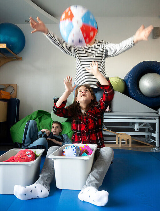 Ein Mädchen und ein Junge sitzen in einem Spielzimmer und greifen nach einem Ball