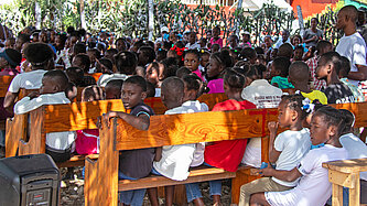 Kinder sitzen auf Holzbänken im Schatten eines großen Baumes.