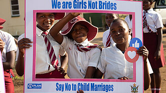 Drei Schülerinnen in Schuluniform halten gemeinsam einen Rahmen mit der Aufschrift „We are Girls Not Brides – Say No to Child Marriages“.