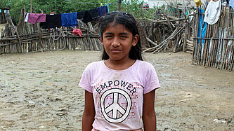 Ein Mädchen steht in einem schlammigen Innenhof und schaut in die Kamera. Hinter ihr hängen Klamotten zum Trocknen über einem Holzzaun.