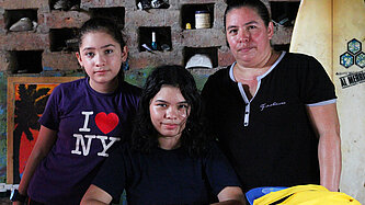 Drei Personen stehen zusammen in einem einfachen Raum mit Tisch und Surfbrett im Hintergrund – Symbolbild für Familien und Jugendliche in einem Projekt für Schutz und Bildung in El Salvador.