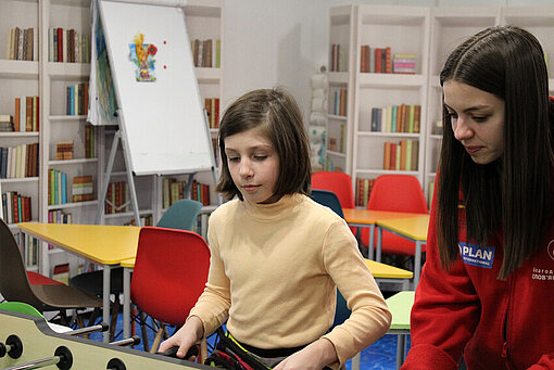Im Kinderschutzraum gibt es für Helga auch einen Tischkicker, an dem sie gerne ihre Kicker-Künste perfektioniert Ein junges Mädchen spielt zusammen mit einer jungen Frau Tischkicker