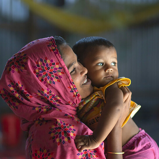 Eine junge Frau mit Kopftuch hält ein Kleinkind auf dem Arm und schaut es lächelnd an