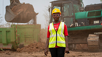 Eine Frau steht in Warnweste und mit Schutzhelm auf einer Baustelle, hinter ihr ist ein Bagger in Aktion
