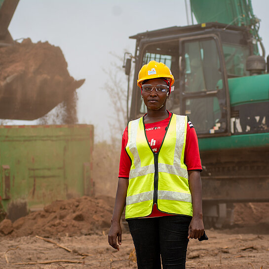 Eine Frau steht in Warnweste und mit Schutzhelm auf einer Baustelle, hinter ihr ist ein Bagger in Aktion