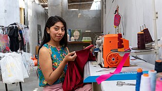 Eine junge Frau sitzt an einer Overlock-Nähmaschine und näht ein Kleidungsstück in ihrer Werkstatt.