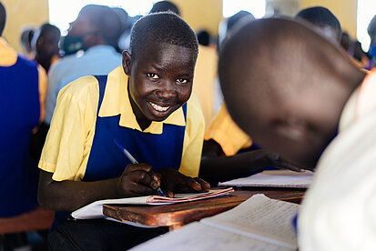 Ein Mädchen sitzt in einem Klassenraum und hält einen Stift über ihrem Heft. Sie schaut in die Kamera und lächelt. 