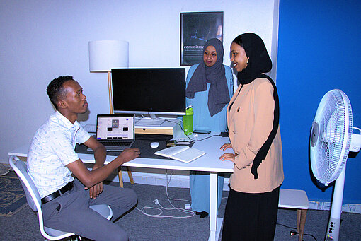 Ein junger Mann sitzt an einem Schreibtisch vor einem Computer. Soumaya steht neben ihm und die beiden unterhalten sich. Hinter dem Schreibtisch steht eine junge Frau, die ebenfalls an der Unterhaltung teilnimmt
