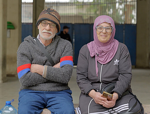 Zwei ältere Erwachsene sitzen nebeneinander auf einer Steinbank. Eine der Personen trägt ein Kopftuch und hält ein Smartphone. 