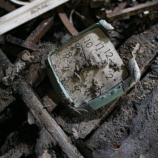 Eine kaputte Wanduhr liegt auf dem Boden, halb von Schutt und Asche verdeckt.